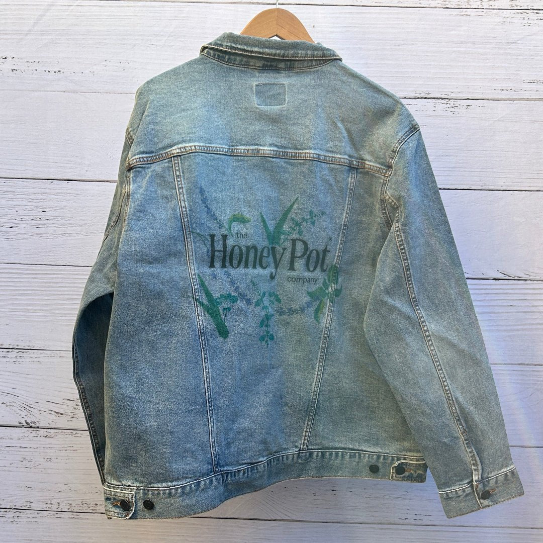 photo of the back artwork on a stonewashed denim jacket showing the logo text “the Honey Pot company” printed in black and white, surrounded by illustrated botanical elements including green leaves, lavender sprigs, aloe-like plants, and small flowering stems arranged in a circular composition; the artwork has clean outlines and soft color fills with visible denim grain and seams framing the print, printed by DenimINK