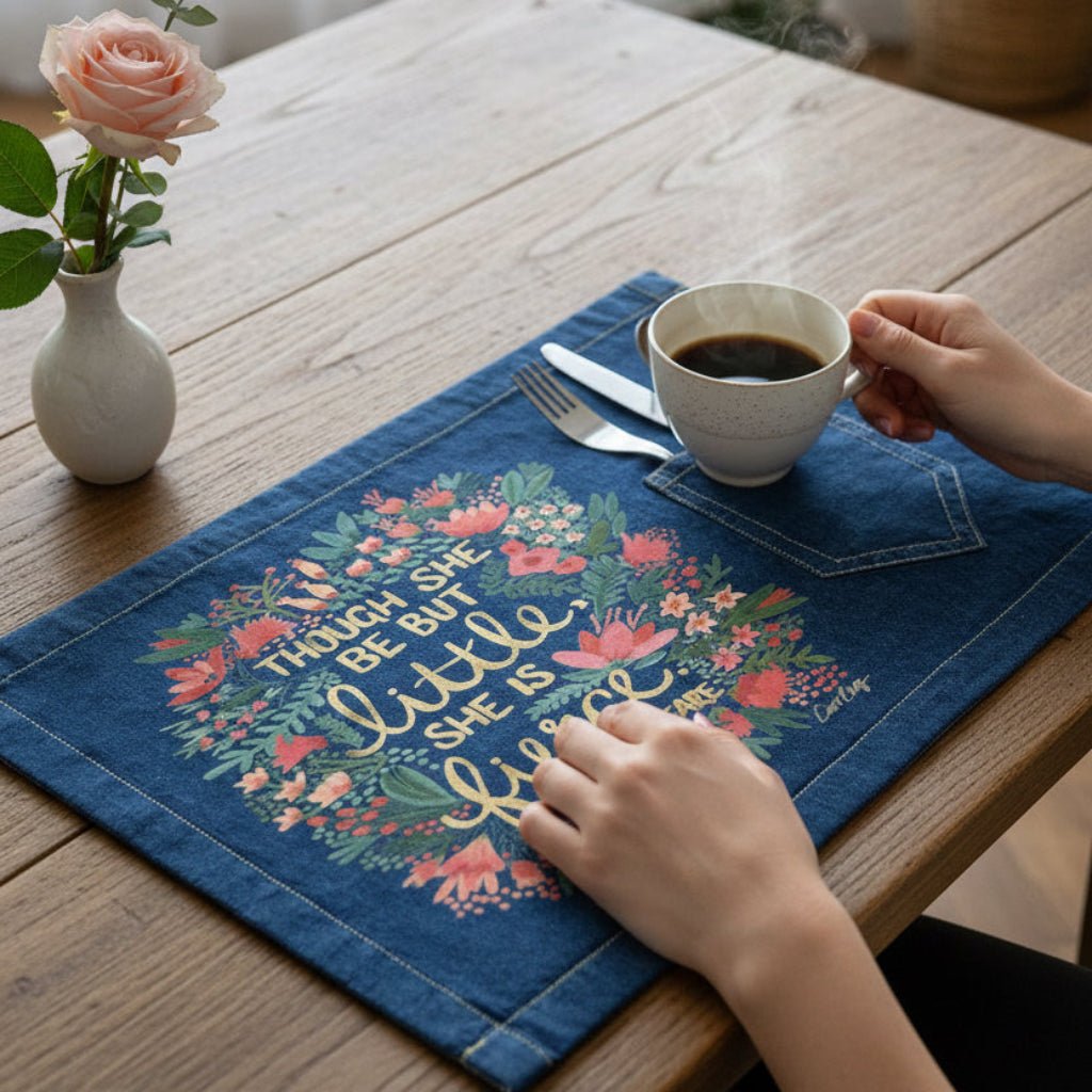 Person holding a cup of coffee on a decorative placemat with floral design and text. with Shakespears quote "Though She Be But Little, She is Fierce" printed by DenimINK. Little and Fierce by CatCoq on a denim table placemat lifestyle shot