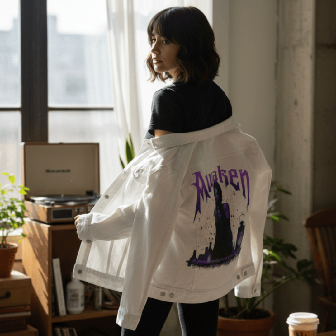 three-quarter view of a model standing near a sunlit window in a softly lit room, holding open a white denim jacket to reveal the printed artwork “Awaken” by DeRouche Designs on the back. the illustration shows a gothic figure in violet hues, surrounded by potion bottles and a lantern beneath dramatic purple text that reads “Awaken.” the artwork is digitally printed on the jacket’s back panel by DenimINK.