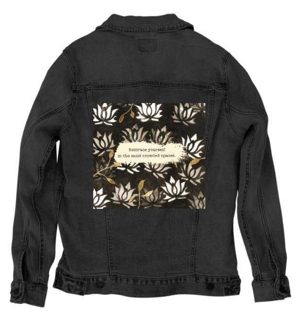 back view of a black denim jacket displaying the artwork “Be Yourself” by Canvas & Quotations, featuring layered white lotus motifs, gold leaf elements, and the quote “Embrace yourself in the most crowded spaces” on a painted cream strip. the artwork is digitally printed on the jacket’s back panel by DenimINK