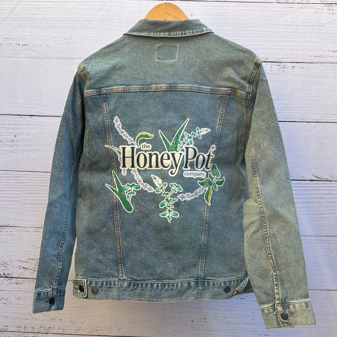 photo of the back artwork on a stonewashed denim jacket showing the logo text “the Honey Pot company” printed in black and white, surrounded by illustrated botanical elements including green leaves, lavender sprigs, aloe-like plants, and small flowering stems arranged in a circular composition; the artwork has clean outlines and soft color fills with visible denim grain and seams framing the print, printed by DenimINK