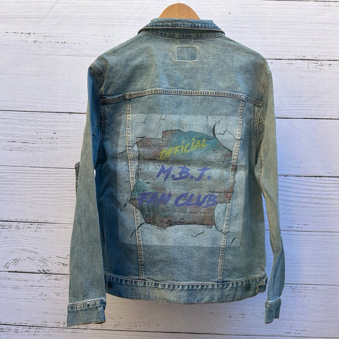 photo of the back artwork on a stonewashed denim jacket showing distressed text reading “OFFICIAL” in yellow and “M.B.J. FAN CLUB” in purple, printed over a textured, weathered surface resembling peeling paint and cracked concrete, with layered gray, teal, and rust tones creating a worn, gritty appearance; the rectangular print is framed by visible denim seams and stitching, printed by DenimINK