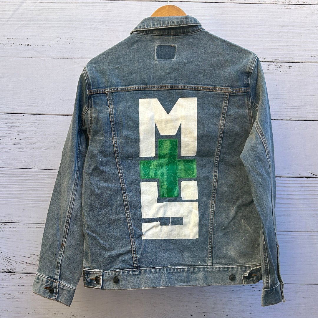 photo of the back artwork on a stonewashed denim jacket showing a bold graphic composed of large white block letterforms arranged vertically around a centered green plus symbol, the plus rendered with a mottled, textured green finish that shows subtle wear and speckling; the graphic is framed by visible denim seams and stitching with natural fabric grain and light distressing throughout, printed by DenimINK