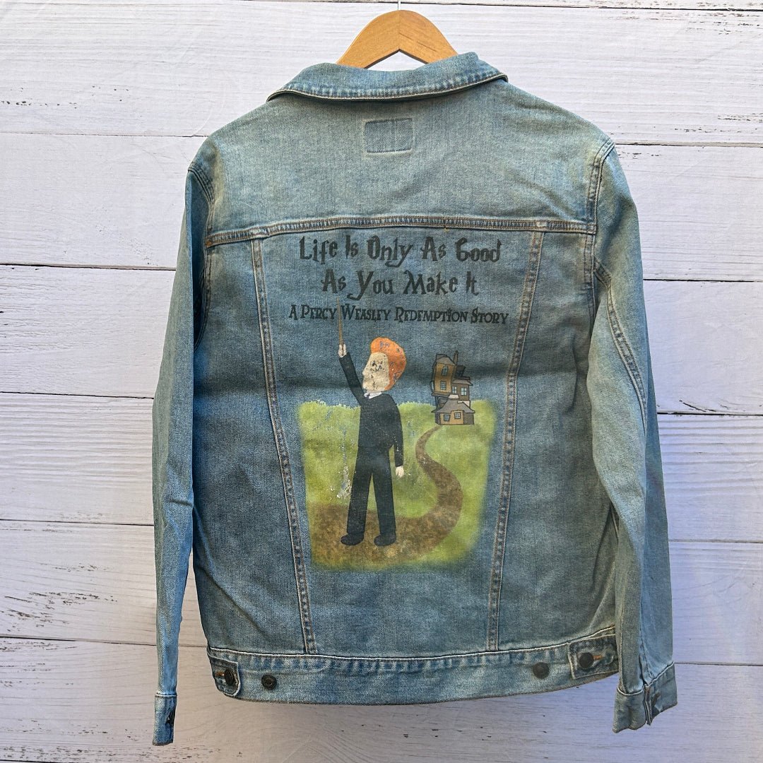 AS - IS. 9. Photo of a stonewashed denim jacket shown from the back on a wooden hanger, featuring a centered illustrated scene beneath hand-lettered text reading “Life Is Only As Good As You Make It” with smaller text reading “A Percy Whiskey Redemption Story,” depicting a red-haired figure in dark clothing standing on a winding dirt path through green grass and pointing upward, with a small house in the distance; the artwork has a storybook, hand-painted style with a softly distressed print texture and vis
