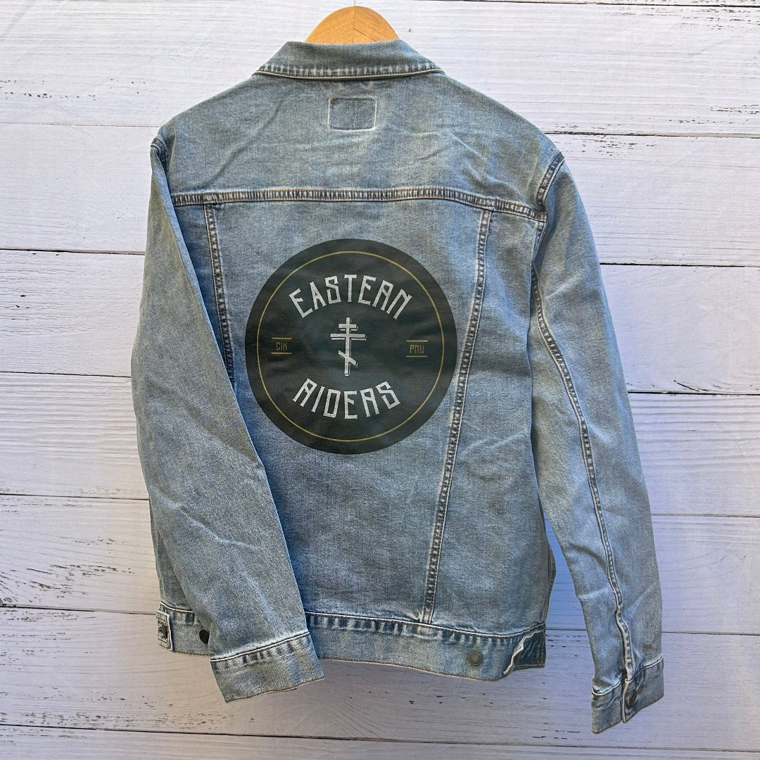 photo of the back artwork on a stonewashed denim jacket showing a circular black emblem printed on the back panel with white arched text reading “EASTERN” at the top and “RIDERS” at the bottom, a white Orthodox-style cross centered inside the circle, thin yellow ring accents, and small yellow text reading “GIK” on the left and “PIW” on the right; the emblem has a slightly worn, matte print texture with visible denim grain and seams framing the artwork, printed by DenimINK