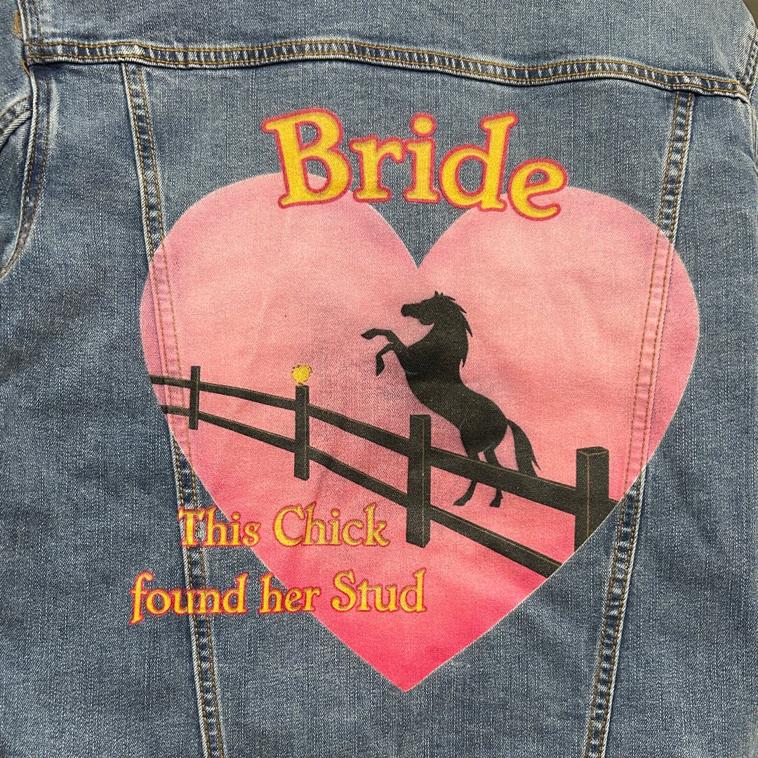closeup Photo of a stonewashed denim jacket shown from the back on a wooden hanger, featuring a large pink heart-shaped illustration with the word “Bride” at the top and the phrase “This Chick found her Stud” below, depicting a black silhouette of a rearing horse behind a fence set against a warm sunset gradient; the artwork has a playful, western-inspired feel with soft, slightly distressed print texture and visible denim grain and seams framing the image, printed by DenimINK