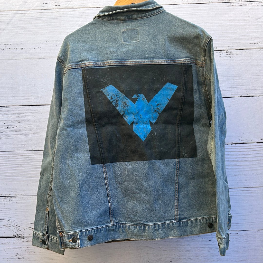 AS - IS. 18. Blue Winged Emblem. ONE - OF - ONE. Printed Denim Jacket — XL / Stonewashed custom denim jacket showing a bold blue winged emblem printed on a black rectangular patch, the emblem formed by sharp angular shapes resembling outstretched wings with a central downward point, rendered in bright blue with intentional distressing, speckling, and worn texture that reveals the fabric beneath; visible denim seams and stitching frame the patch area, printed by DenimINK