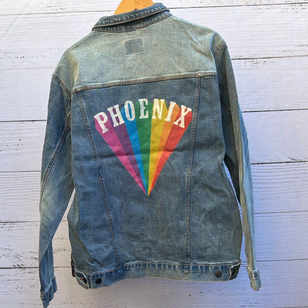 AS - IS. 19. Phoenix Rainbow Rays ONE - OF - ONE. Printed Denim Jacket. Large / Stonewashed. custom denim jacket showing the word “PHOENIX” printed in distressed white block letters arched above a fan of rainbow-colored rays, with vertical bands of pink, purple, blue, green, yellow, orange, and red converging to a point near the bottom of the back panel; the print has a slightly worn, textured finish with visible denim grain and seams surrounding the artwork, printed by DenimINK