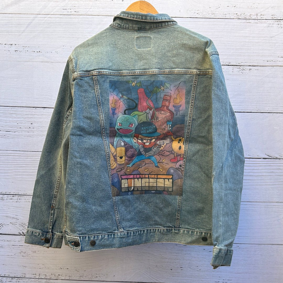 AS - IS. 20. Enter The Gungeon. ONE - OF - ONE. Printed Denim Jacket. XL / Stonewashed. custom denim jacket showing a colorful cartoon-style dungeon scene with a bearded character wearing a cap reading “KAMIKAZE WAS WRONG” holding a gun, surrounded by bullet-shaped characters, fantasy creatures, torches, bottles, and scattered shell casings inside a stone-walled dungeon; visible text at the bottom of the illustration reads “ENTER THE GUNGEON,” with vibrant blues, reds, greens, and yellows printed within a r