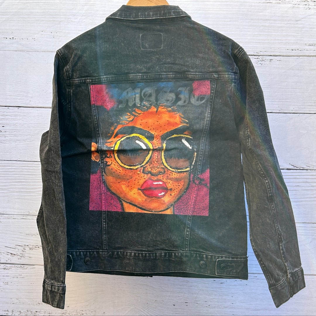 photo of the back artwork on a black denim jacket showing a painted portrait of a woman with warm orange skin tones, freckles, glossy red lips, and oversized round yellow glasses with dark lenses, set against a magenta and deep blue background; the illustration has bold outlines, visible brush texture, and a slightly distressed print finish with denim grain and seams framing the rectangular artwork, printed by DenimINK