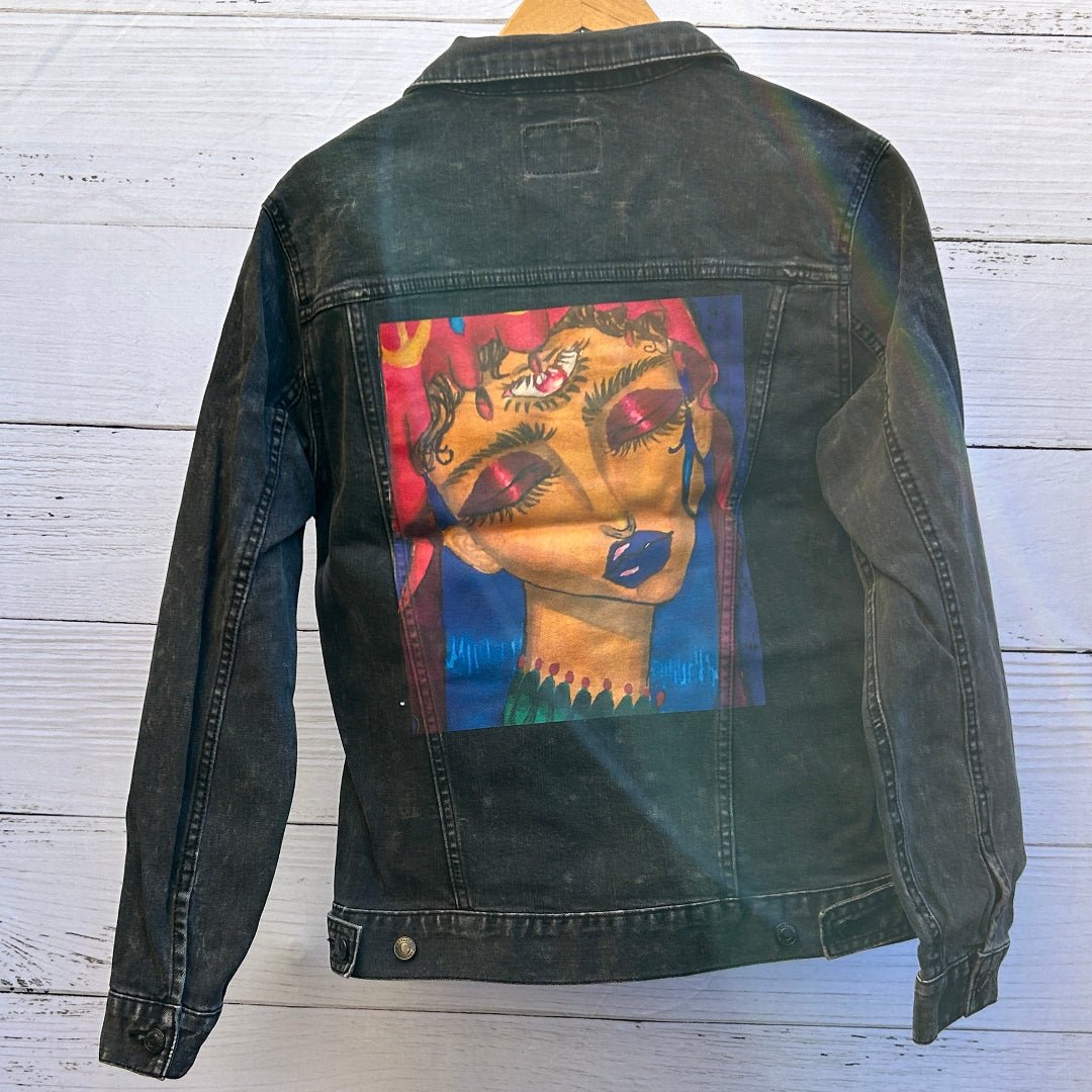 photo of the back artwork on a black denim jacket showing a richly painted portrait of a woman with closed eyes, deep red eyelids, long lashes, blue lips, and warm golden skin tones, rendered with expressive brushstrokes in red, blue, teal, and pink within a rectangular print area; the artwork has a soft, painterly texture with visible fabric grain and denim seams framing the image, printed by DenimINK