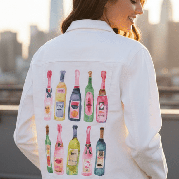 three-quarter rear view of a smiling model standing on a rooftop with a distant city skyline and skyscrapers blurred in the background. they wear a white denim jacket printed with the “Champagne Collection” artwork by CatCoq on the back panel. the illustration features twelve watercolor champagne bottles in two rows, each in different pastel and jewel tones with playful label designs. soft golden-hour light reflects off the white fabric, enhancing the clarity and color vibrancy of the printed design.