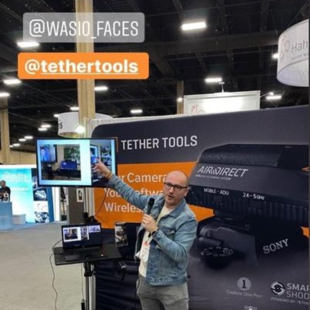 Back view of a photographer wearing a stonewashed denim jacket branded with “WASIO Photography” while presenting a live demo at a busy trade show booth. Screens glow with tethered images, cameras and cables frame the space, and the jacket stands out as a confident marker of identity amid the buzz of gear, conversation, and shared learning.