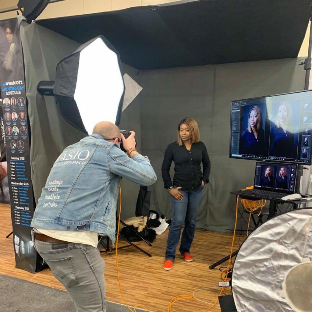 Back view of a photographer actively shooting a portrait in a studio environment while wearing a stonewashed denim jacket branded with “WASIO Photography.” Softboxes glow, cables snake across the floor, and the jacket’s printed service list is visible as the photographer leans into the moment. The scene hums with focus and motion, blending craft, branding, and real-time creativity. Printed by DenimINK