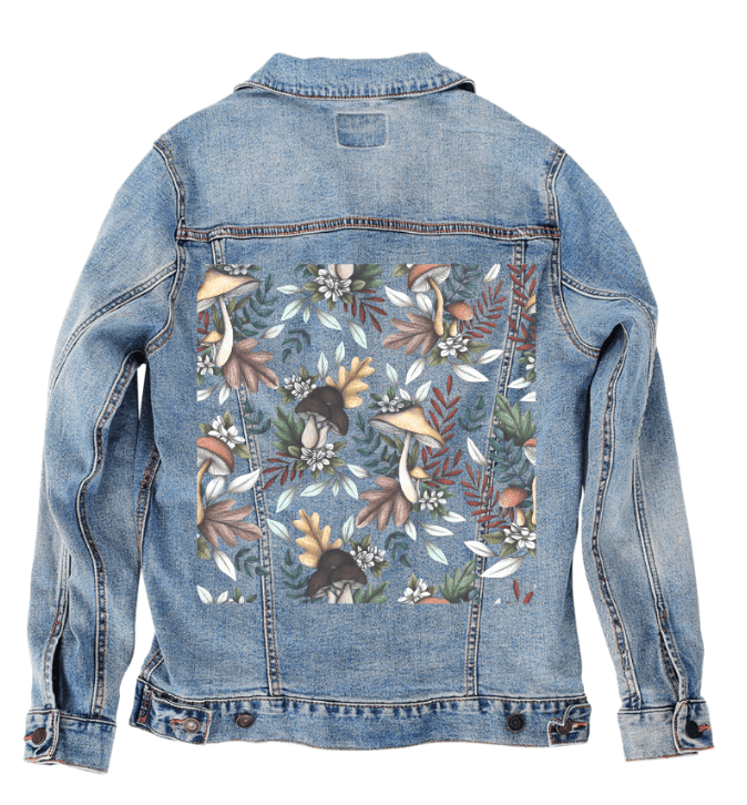 back view of a stonewashed denim jacket featuring the artwork “Enchanted Mushrooms” by Cecilia Battaini, a soft, painterly repeat of mushrooms in amber, wine & charcoal tones surrounded by fern fronds, pinecones, clover & botanical textures. the gentle fade of the denim creates the effect of walking through a sun-dappled woodland trail. the design is digitally printed on the jacket’s back panel by DenimINK.
