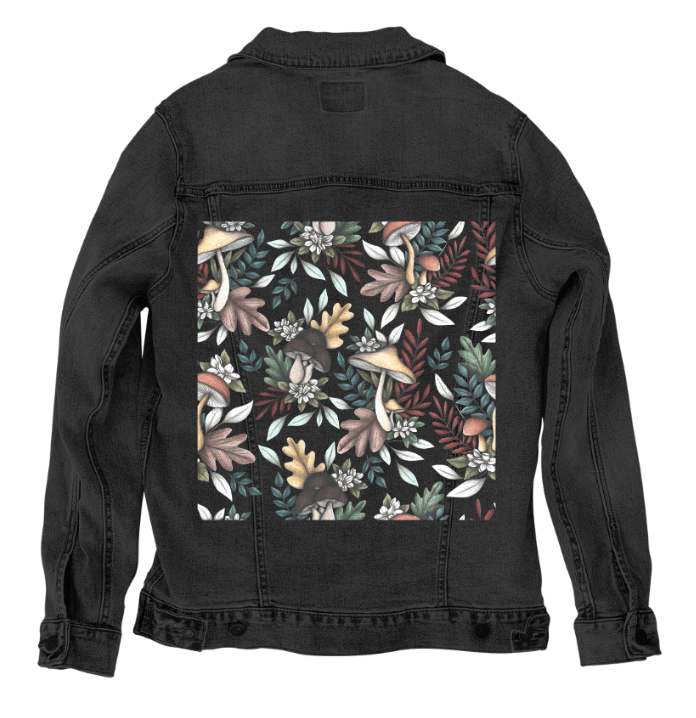 back view of a black denim jacket featuring the artwork “Enchanted Mushrooms” by Cecilia Battaini, a richly detailed pattern of forest mushrooms, fern tendrils, fallen oak leaves & tiny ivory flowers. bold shapes in burgundy, ochre & moss green bloom against the dark fabric like forest life emerging from shadow. the design is digitally printed on the jacket’s back panel by DenimINK.