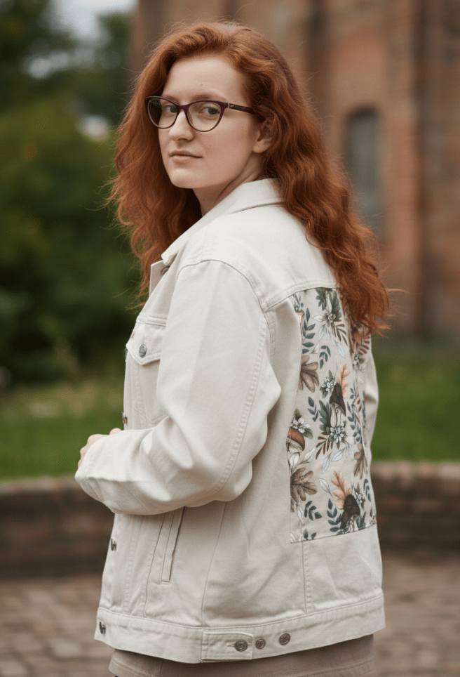 back view of a model standing near a weathered red brick wall wearing a white denim jacket featuring the artwork “Enchanted Mushrooms” by Cecilia Battaini, a clean, delicate composition of illustrated mushrooms, clovers, and pale forest ferns in soft reds, greens, greys & creams. each line feels etched from nature, like pressed leaves in an old botanical notebook. the design is digitally printed on the jacket’s back panel by DenimINK.