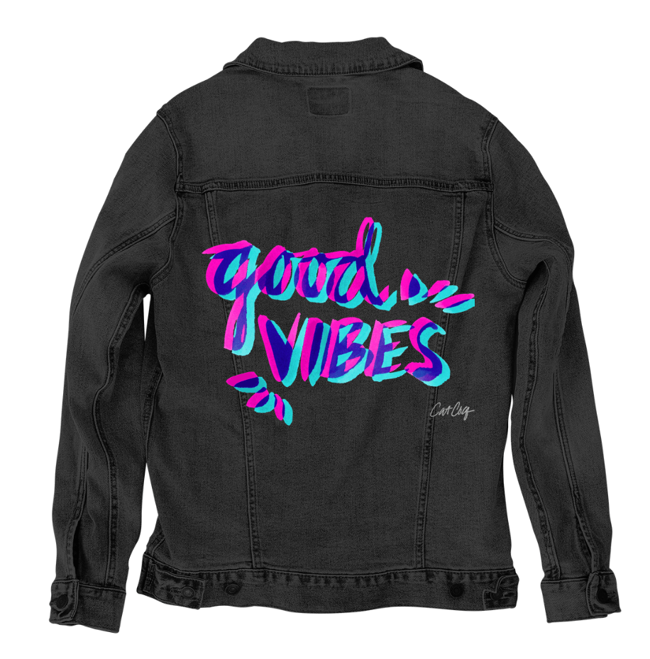 back view of a black denim jacket featuring the artwork “Good Vibes” by CatCoq, showing overlapping hand-lettered brush strokes in bright pink, teal, and blue. the vibrant colors contrast sharply with the black denim fabric, making the lettering stand out with a neon-like effect. the artwork is digitally printed on the jacket’s back panel by DenimINK.