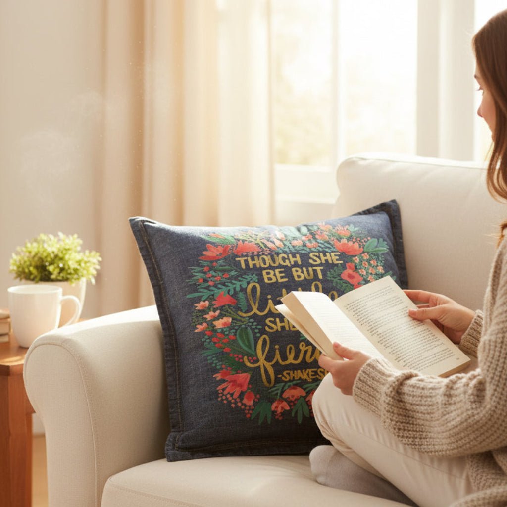 Person reading a book on a couch with a decorative denim pillow with Shakespears quote "Though She Be But Little, She is Fierce" printed by DenimINK. Little and Fierce by CatCoq on a denim throw pillow lifestyle shot