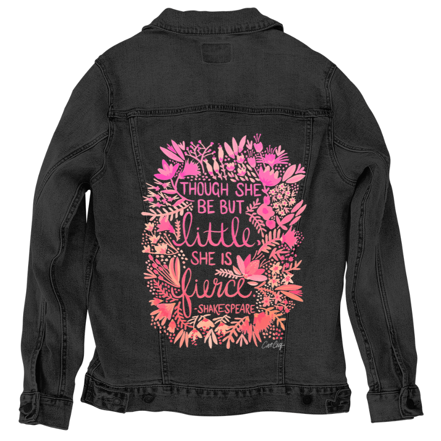 back view of a black denim jacket featuring the “Little & Fierce” artwork by CatCoq, digitally printed by DenimINK. the quote “Though she be but little, she is fierce” appears in yellow lettering surrounded by an oval-shaped floral frame. the detailed wreath includes layered leaves & blooms in warm pinks, coral, sage & green. the dark jacket fabric enhances the vibrant colors of the watercolor design.