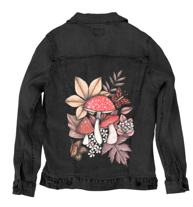 back view of a black denim jacket featuring a printed illustration of red mushrooms with speckled caps, surrounded by shaded forest leaves, blossoms, & butterflies in deep ochre & rust tones. the artwork “Mushroom Magic 1” by Cecilia Battaini is digitally printed on the jacket’s back panel by DenimINK.
