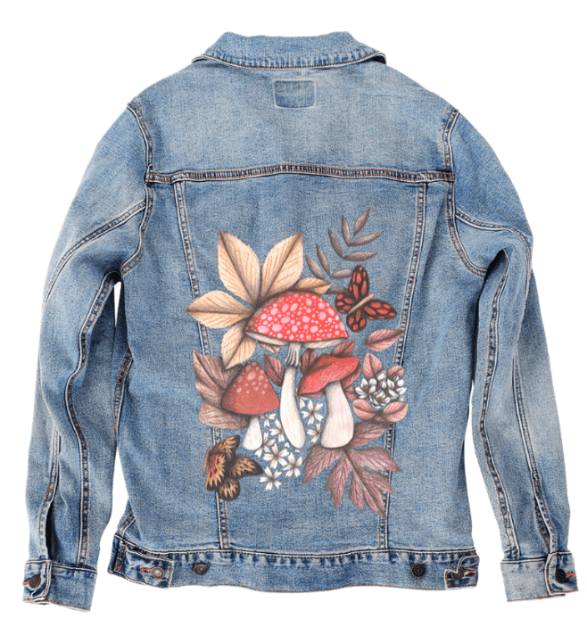 back view of a stonewashed denim jacket featuring a printed cluster of red-capped mushrooms surrounded by soft fallen leaves, white blossoms, & two butterflies in flight. the artwork “Mushroom Magic 1” by Cecilia Battaini is digitally printed on the jacket’s back panel by DenimINK.