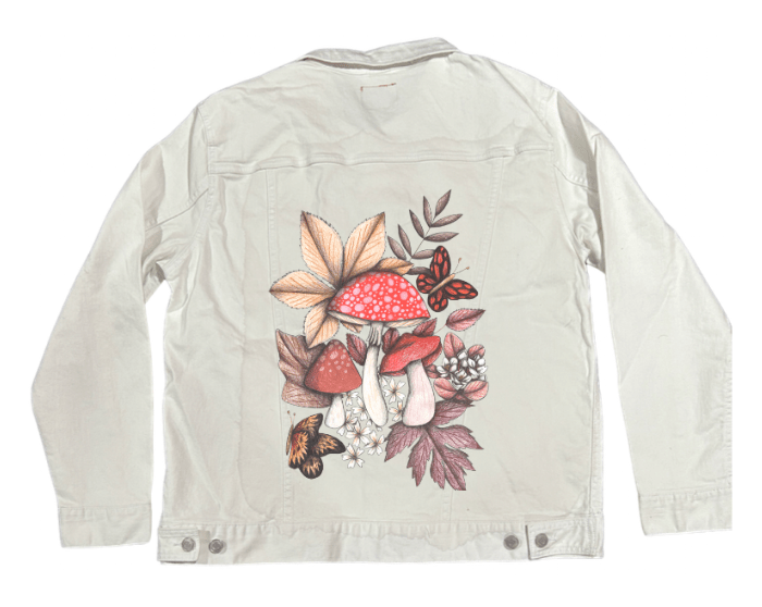 back view of a white denim jacket featuring a printed cluster of woodland mushrooms with red caps, delicate pencil-shaded stems, and scattered blossoms, framed by light leaves and two butterflies in soft motion. the artwork “Mushroom Magic 1” by Cecilia Battaini is digitally printed on the jacket’s back panel by DenimINK.