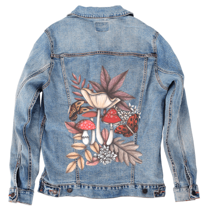 back view of a stonewashed denim jacket featuring a hand-drawn cluster of forest elements — red toadstools, golden butterflies, sepia leaves & soft white blooms. the artwork “Mushroom Magic 3” by Cecilia Battaini is digitally printed on the jacket’s back panel by DenimINK.