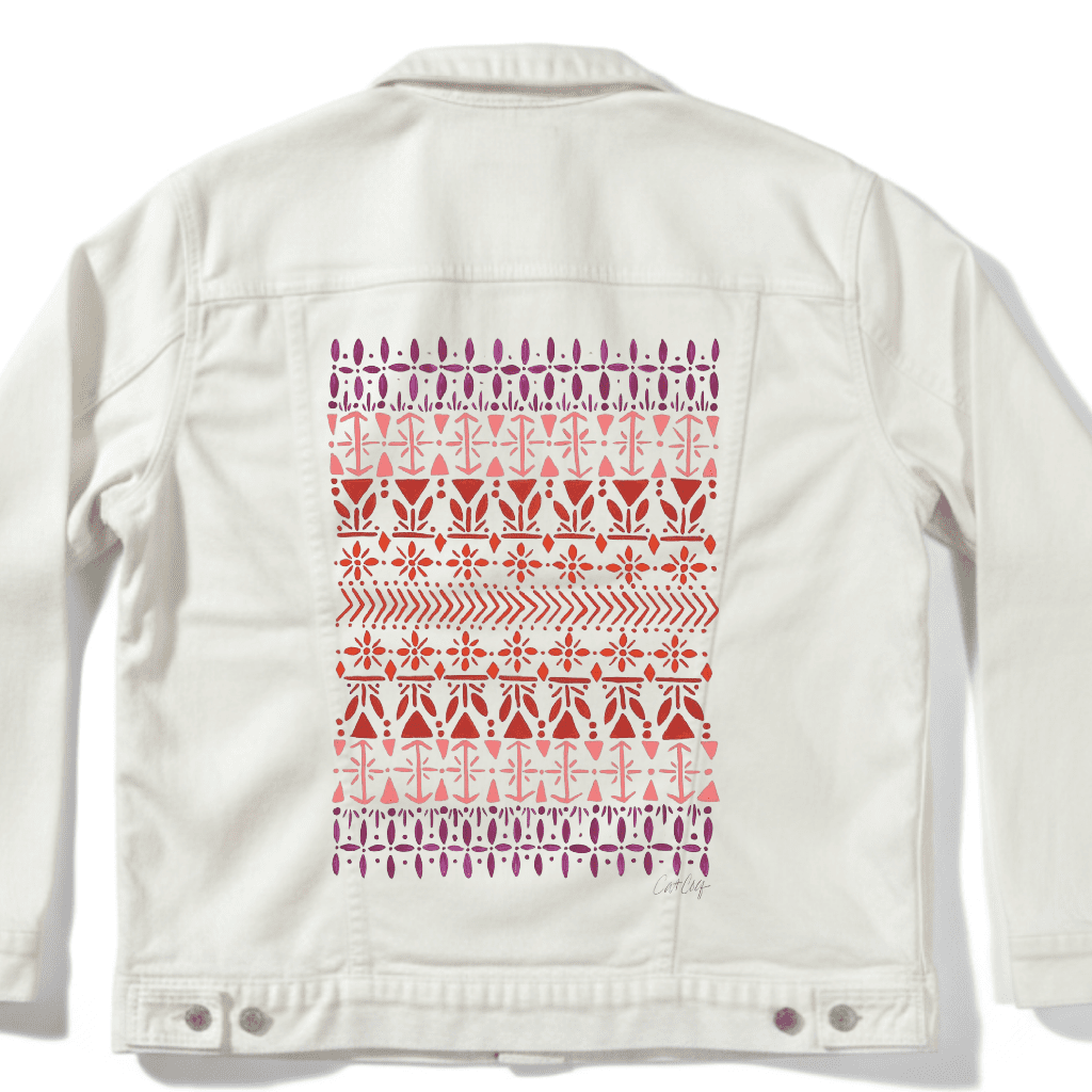White denim jacket with a vertically aligned Scandinavian folk pattern "Norwegian Pattern" by CatCoq made of repeating geometric rows including tulip shapes, arrows, diamonds, snowflakes & stylized trees. the hand-painted elements fade from deep violet at the top to coral & rust red toward the center, then shift to pink & purple again. printed by DenimINK