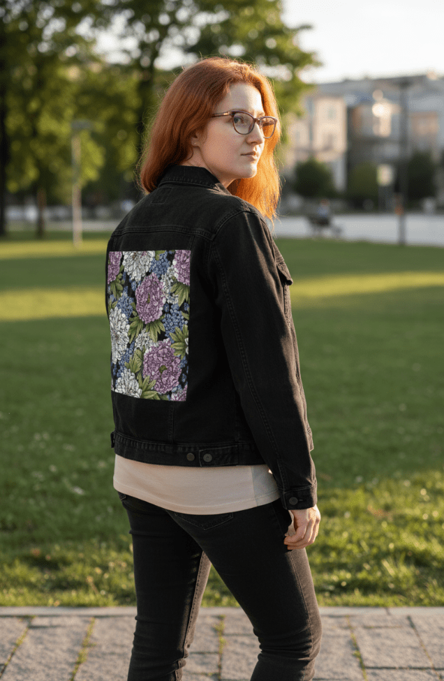 three-quarter rear view of a model standing in the golden light of late afternoon at a city park, wearing a black denim jacket featuring a bold printed panel of white and lavender peonies layered with green leaves and soft blue wildflowers. the artwork “Peonies” by Cecilia Battaini is digitally printed on the jacket’s back panel by DenimINK.