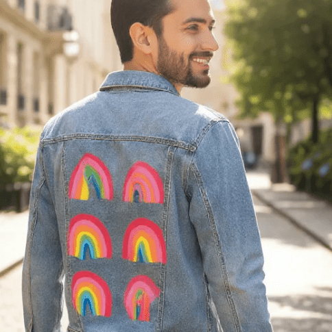 three-quarter rear view of a smiling model on a cobblestone street wearing a stonewashed denim jacket printed with the “Rainbow Collection” by CatCoq. the design shows six hand-painted rainbows arranged in two columns, each with vibrant arcs of red, orange, yellow, green, blue & purple. sunlight highlights the texture of the watercolor artwork. printed by DenimINK.