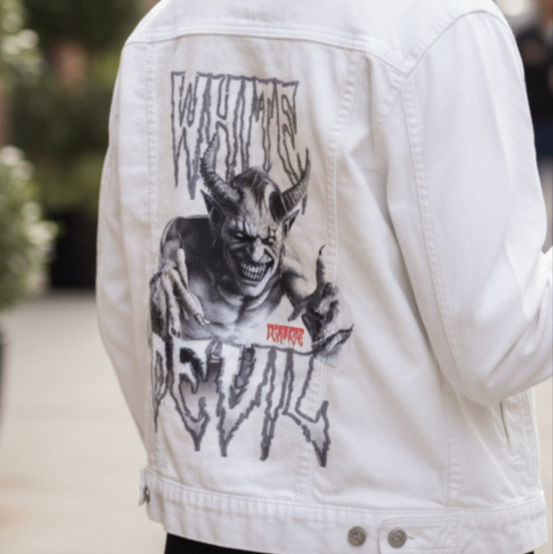 Close-up of a white denim jacket worn by a person walking outdoors on a bright day featuring an illustration of a menacing horned demon creature lunging forward with outstretched clawed hands in an aggressive pose. The demon features prominent curved horns. WHITE DEVIL by DeRouche Designs, printed by DenimINK.
The camera focuses on the jacket's back, capturing crisp fabric texture and contrast stitching.