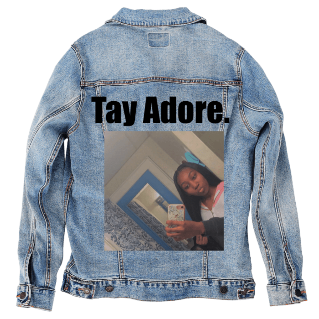 Customer designed custom denim jacket (S stonewash) featuring bold black lettering reading “Tay Adore.” across the upper back. Below it, a softly lit mirror selfie shows a young woman holding a patterned phone case, standing in a bedroom with blue-framed doors, textured curtains, and pale walls. Her long hair and hoodie catch gentle indoor light, creating warm highlights. The calm everyday setting contrasts with the rugged stonewash denim, printed on denim jacket by DenimINK.