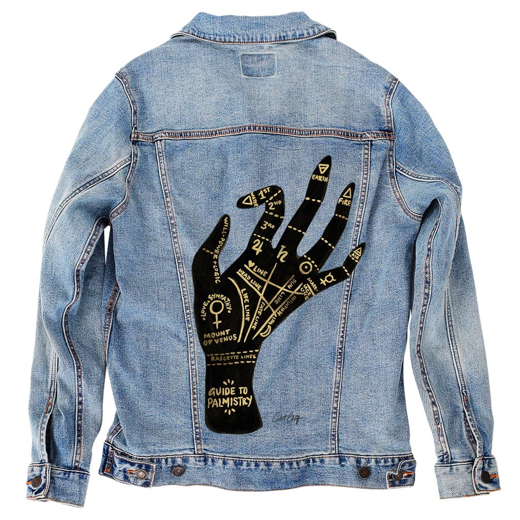back view of a stonewashed denim jacket featuring the “Palmistry” artwork by CatCoq. the design shows a black palm with golden linework outlining classic palmistry zones—heart, life, fate, & mounts—surrounded by astrological & elemental symbols. the soft denim wash gives the design a worn-in, vintage look while preserving the mystical detail. printed by DenimINK.