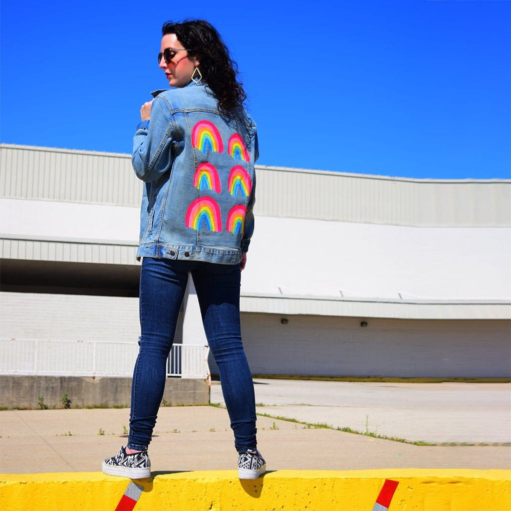 full rear view of a model standing on a yellow painted curb against a vivid blue sky, wearing an oversized stonewashed denim jacket printed with the “Rainbow Collection” by CatCoq. the bold brush-painted rainbows are arranged in a vertical grid, each in red, yellow, green, blue & purple. high-contrast setting gives the artwork a vibrant, pop-art feel. printed by DenimINK.
