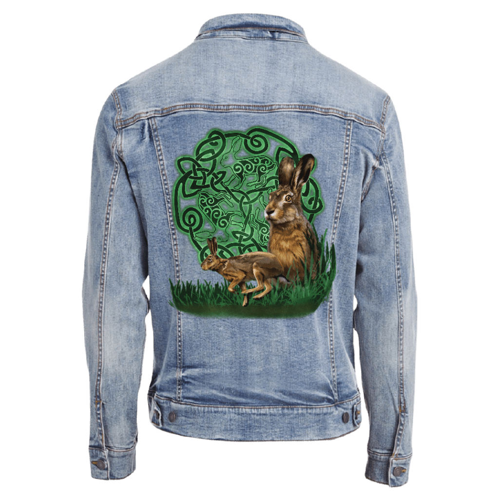 back view of a stonewash blue denim jacket featuring the artwork “celtic hare” by brigid ashwood, printed centered on the back panel. the artwork shows two brown hares, one standing alert and one crouched, positioned in green grass in front of a glowing green celtic knotwork circle with animal motifs woven into the design, clearly visible against the stonewash denim. digitally printed by DenimINK.
