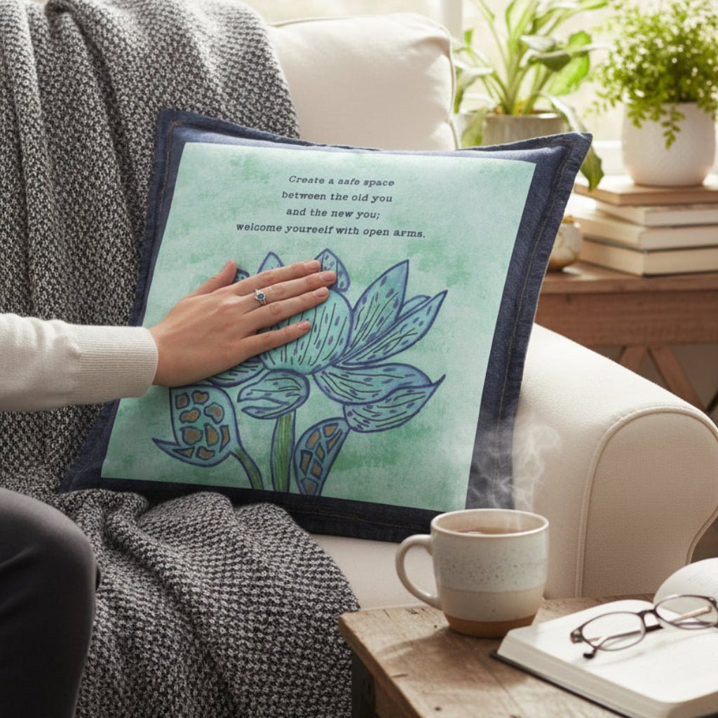 Person holding a decorative denim pillow with a plant design in a cozy living room.  featuring a blue lotus flower on a textured light-green background, paired with the quote “Create a safe space between the old you and the new you; welcome yourself with open arms.” the artwork is titled “Always Welcome Yourself” and created by Canvas &amp; Quotations (Monika Chugh and Alka Chopra) and printed by DenimINK