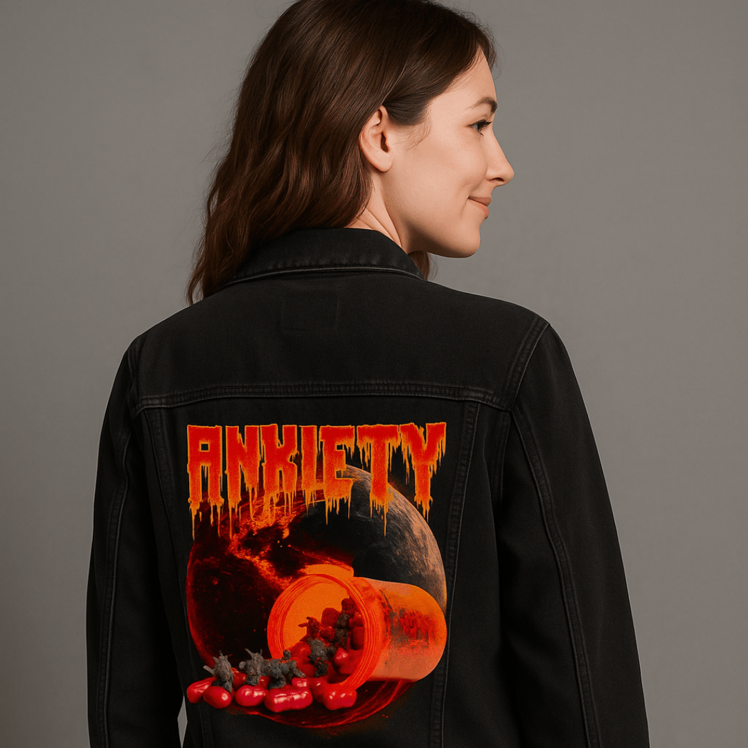 three-quarter rear view of a model posing in a shadowy indoor studio wearing a black denim jacket featuring the artwork “Anxiety” by DeRouche Designs. the design shows red capsules and small devilish figures spilling from a bright orange prescription bottle in front of a scorched, red-lit planet. above, the word “Anxiety” drips in vivid orange and red. the artwork “Anxiety” by DeRouche Designs is digitally printed on the jacket’s back panel by DenimINK.