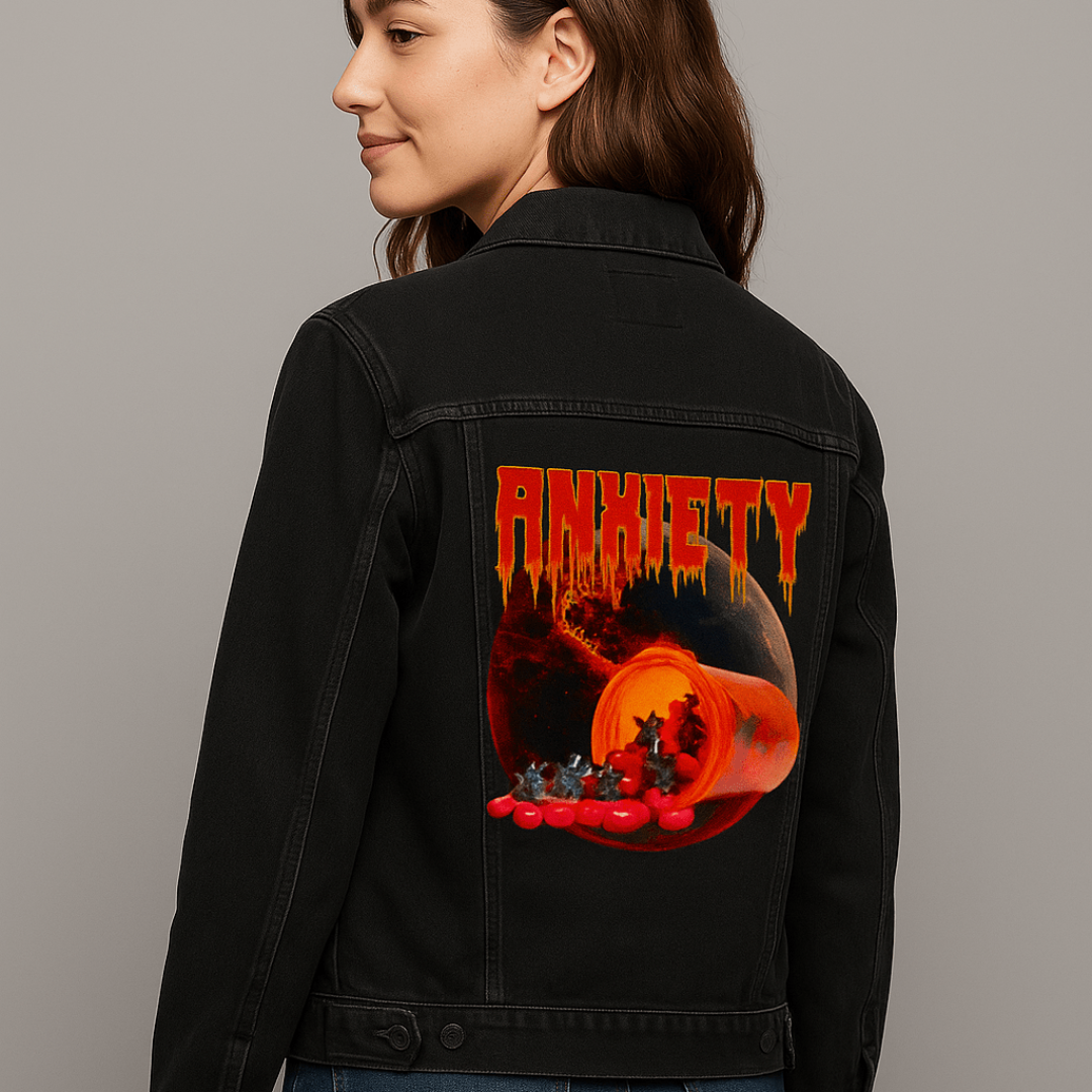 three-quarter view of a model in a neutral indoor studio wearing a black denim jacket featuring the artwork “Anxiety” by DeRouche Designs. the design shows a glowing orange prescription bottle spilling out red capsules and tiny devil figures, set against a burning, cracked planetary background with the word “Anxiety” melting in jagged red text. the artwork “Anxiety” by DeRouche Designs is digitally printed on the jacket’s back panel by DenimINK.