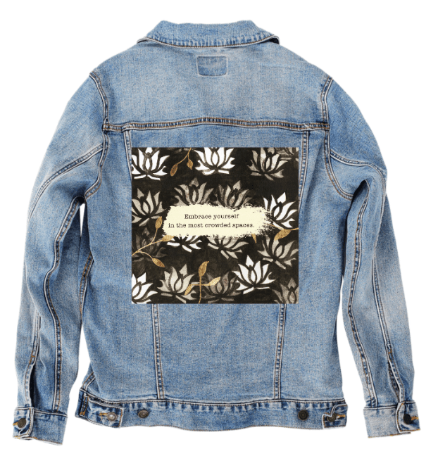 back view of a stonewash denim jacket featuring the artwork “Be Yourself” by Canvas &amp; Quotations, depicting repeating white lotus designs with gold accents and the quote “Embrace yourself in the most crowded spaces.” the artwork is digitally printed on the jacket’s back panel by DenimINK