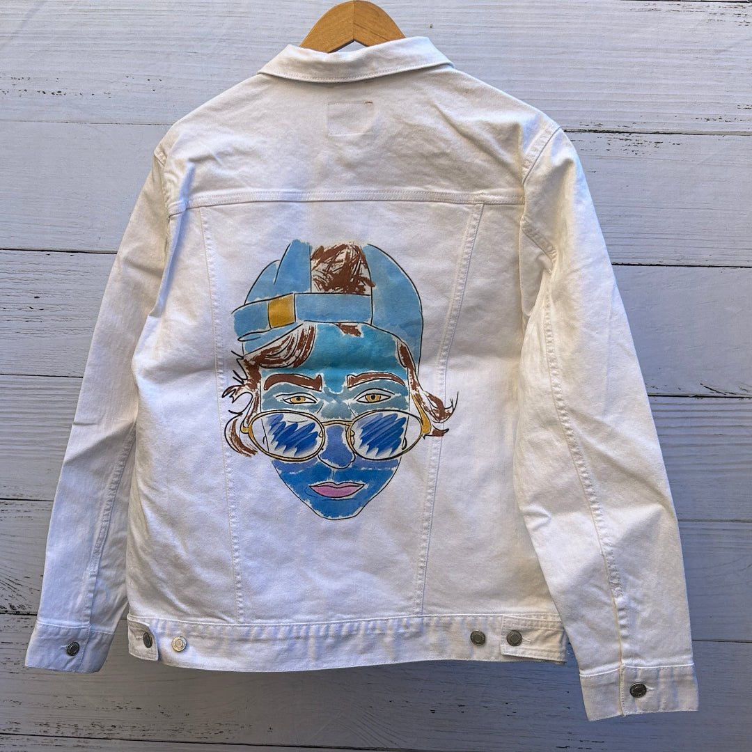 photo of the back artwork on a white denim jacket showing a hand-drawn illustrated face with blue-toned skin, yellow eyes, pink lips, and loose sketch-style lines, wearing a backward blue cap and round gold-rimmed sunglasses filled with scribbled blue shading; brown hair peeks out beneath the cap, and the illustration has a watercolor-and-ink texture with visible brush strokes and denim seams framing the artwork, printed by DenimINK