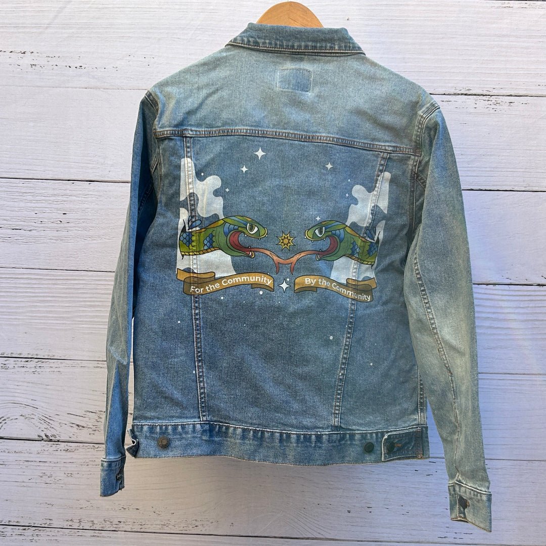 photo of the back artwork on a stonewashed denim jacket showing two mirrored illustrated green snakes facing each other with red tongues extended toward a small yellow starburst at the center, surrounded by white cloud-like shapes and scattered stars; gold ribbon banners beneath the snakes read “For the Community” on the left and “By the Community” on the right, with clean linework, saturated colors, and visible denim grain and seams framing the artwork, printed by DenimINK
