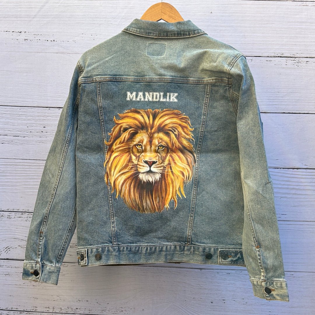 photo of the back artwork on a stonewashed denim jacket showing a highly detailed painted portrait of a lion’s face with piercing golden eyes and a full flowing mane rendered in layered shades of gold, amber, and brown, centered on the back panel beneath white block lettering reading “MANDLIK”; the artwork has realistic fur texture and painterly depth, with visible denim grain and seams framing the print, printed by DenimINK