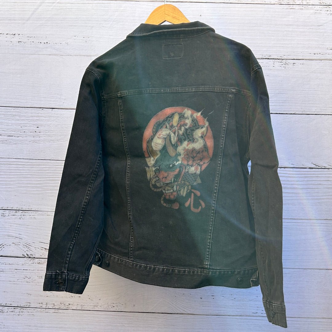 photo of the back artwork on a black denim jacket showing a detailed illustrated skull composed of layered elements including horns, teeth, rope, fabric textures, and organic shapes, rendered in muted red, teal, cream, and brown tones within a rounded composition; a red circular backdrop frames the skull, with a small red character symbol printed near the lower right of the artwork, and the print appears intentionally distressed with visible denim grain and seams surrounding the image, printed by D