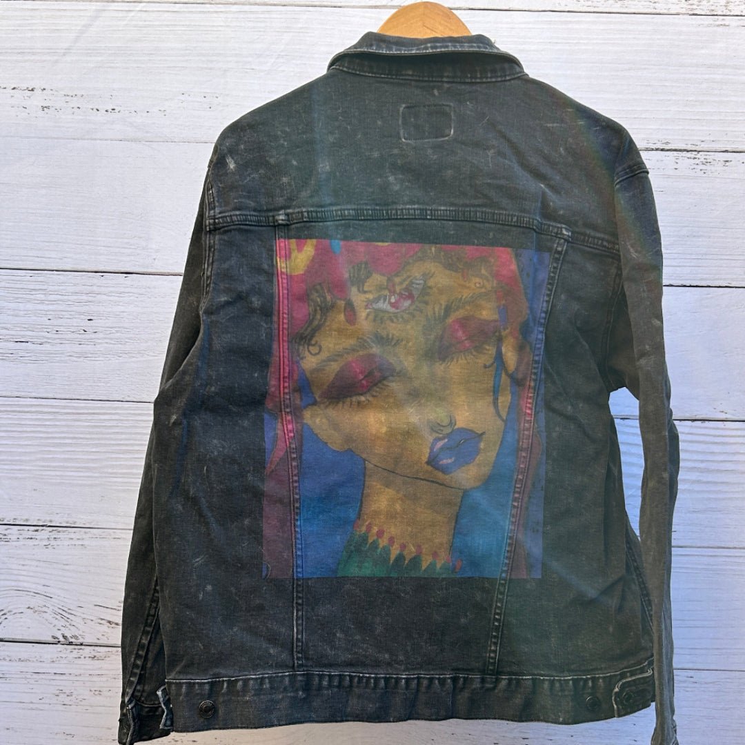 photo of the back artwork on a black denim jacket showing a painted portrait of a woman with closed eyes, long lashes, deep red eyelids, blue lips, and warm golden skin tones, rendered in expressive brushstrokes with pink, blue, and teal accents forming a rectangular composition; the artwork has a soft, painterly texture with slight translucency where the denim grain shows through, and visible seams frame the print, printed by DenimINK