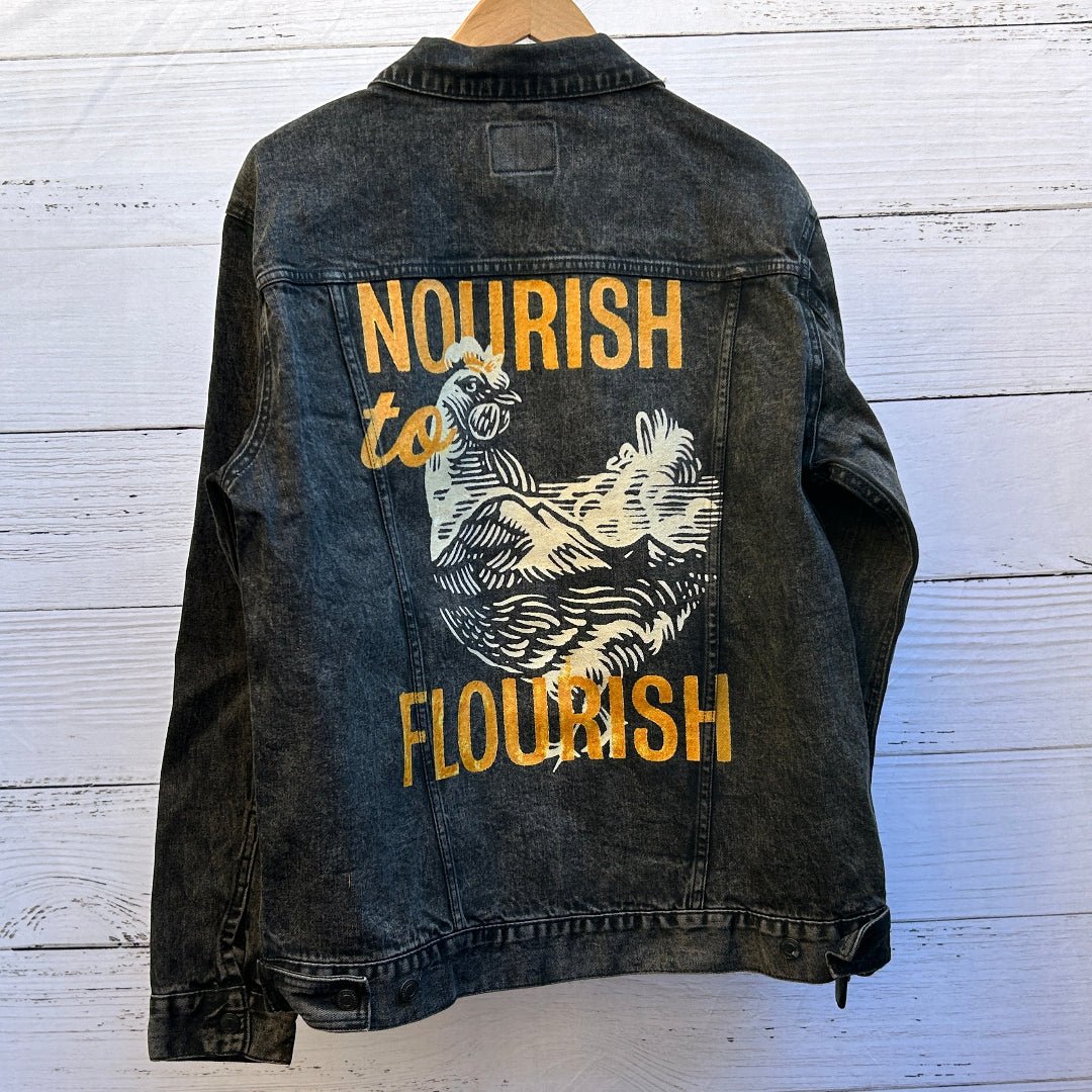 AS - IS. 12. Nourish To Flourish. ONE - OF - ONE. Printed Denim Jacket — Medium / Black custom denim jacket by DenimINK showing a bold linocut-style illustration of a hen rendered in thick white carved lines, surrounded by textured mustard-yellow lettering reading “NOURISH” at the top, “to” partially overlapping the hen’s head, and “FLOURISH” at the bottom; the artwork has a handcrafted, slightly distressed ink texture against the dark denim, with visible weave and seams framing the print