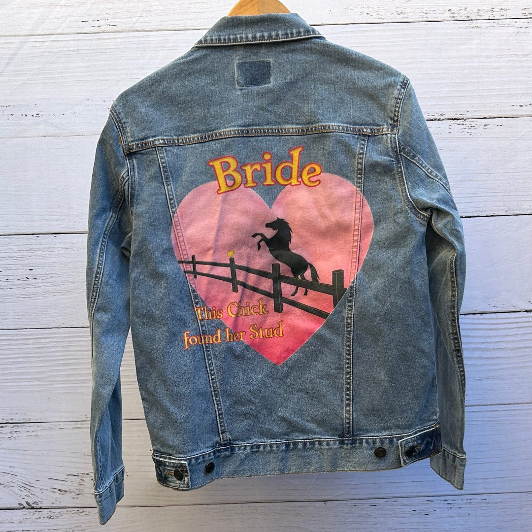 AS - IS. 13. Photo of a stonewashed denim jacket shown from the back on a wooden hanger, featuring a large pink heart-shaped illustration with the word “Bride” at the top and the phrase “This Chick found her Stud” below, depicting a black silhouette of a rearing horse behind a fence set against a warm sunset gradient; the artwork has a playful, western-inspired feel with soft, slightly distressed print texture and visible denim grain and seams framing the image, printed by DenimINK