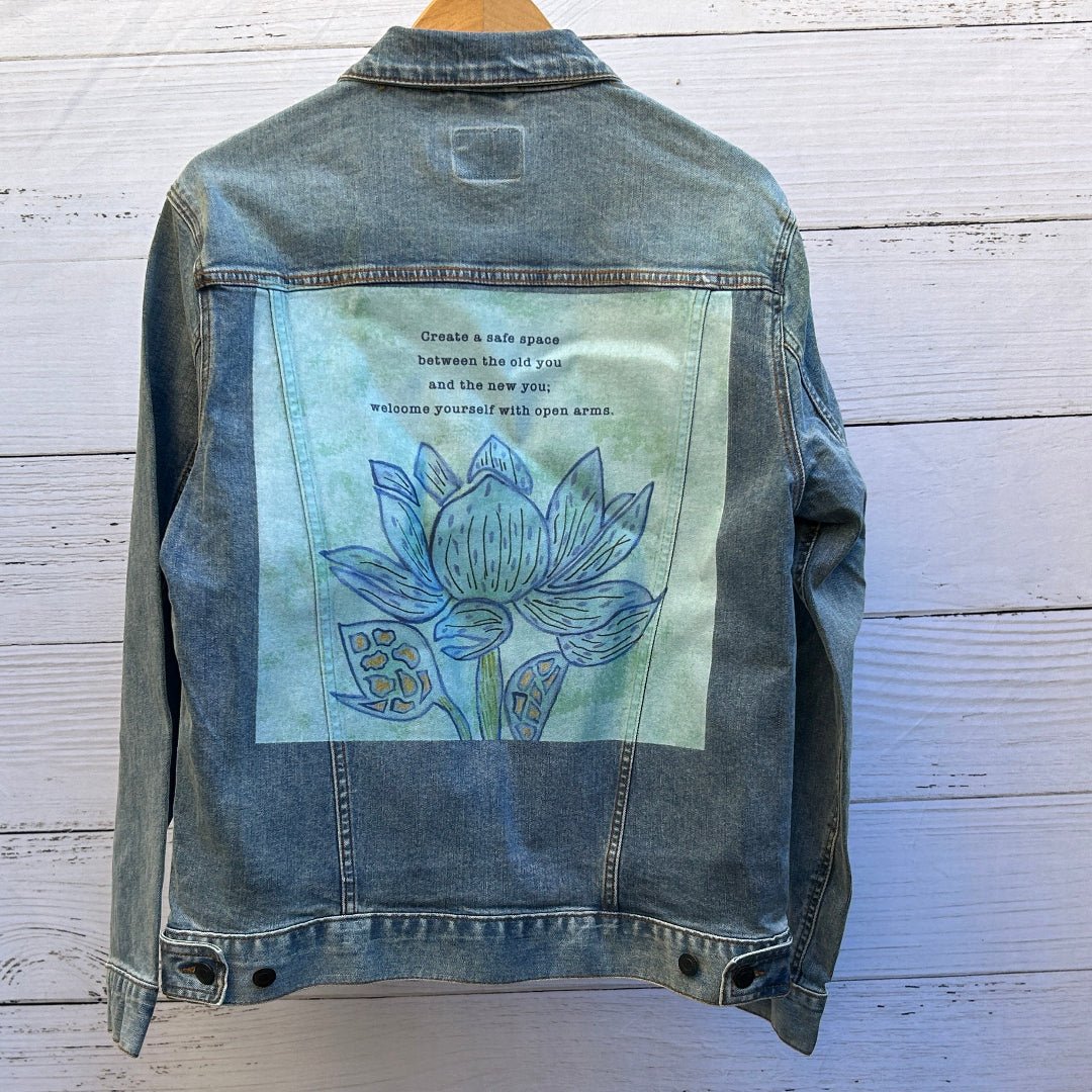 AS - IS. 14. Photo of a stonewashed denim jacket shown from the back, featuring a centered illustrated artwork titled “Always Welcome Yourself” by Canvas &amp; Quotations, depicting a hand-drawn blue lotus flower with layered petals and organic linework set against a soft green, watercolor-style background; above the flower is visible text reading “Create a safe space between the old you and the new you, welcome yourself with open arms,” with a calm, reflective tone and lightly distressed print texture that all
