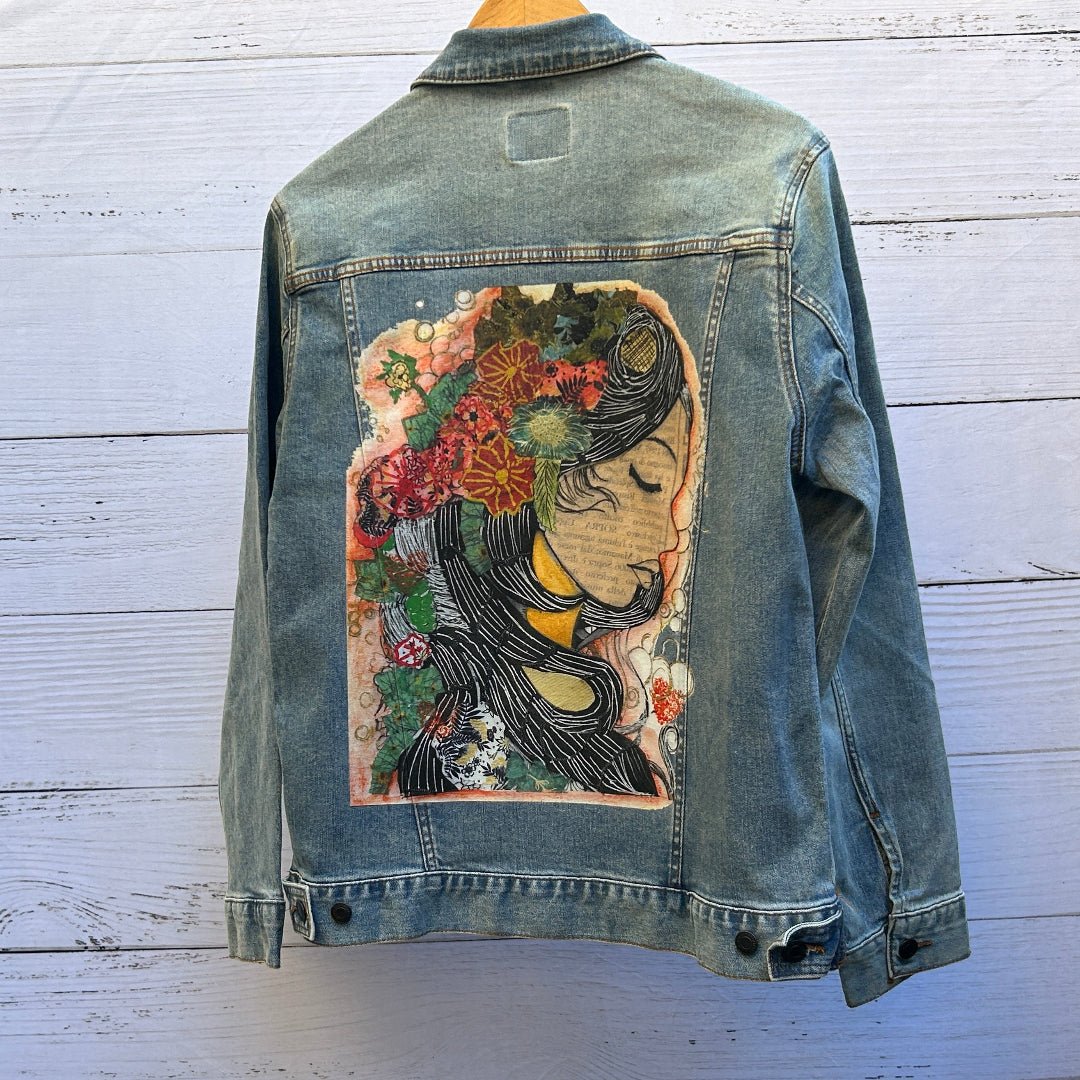 AS - IS. 16. Photo of a stonewashed denim jacket shown from the back, featuring a centered mixed-media collage artwork titled “Coloring Page Woman Collage” by Canvas &amp; Quotations, depicting a serene female profile with closed eyes rendered in layered ink lines, paper textures, and handwritten text fragments, crowned with an intricate arrangement of flowers, leaves, and abstract shapes in warm reds, greens, creams, and blacks; the artwork has a hand-cut, illustrative collage feel with visible texture and sof