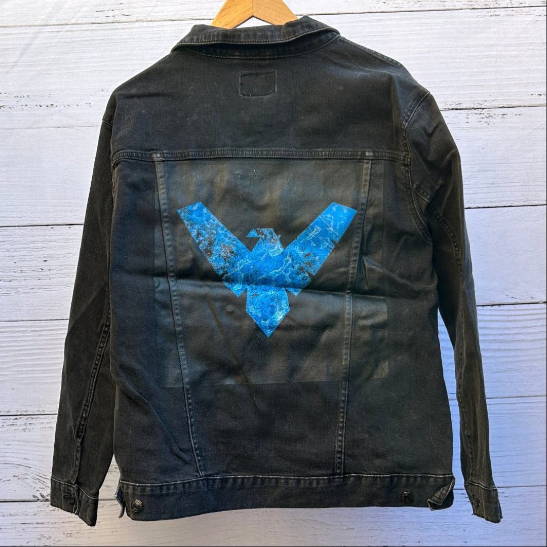 AS - IS. 17. Blue Winged Emblem. ONE - OF - ONE. Printed Denim Jacket — XL / Black custom denim jacket showing a bold blue winged emblem printed on a black rectangular patch, the emblem formed by sharp angular shapes resembling outstretched wings with a central downward point, rendered in bright blue with intentional distressing, speckling, and worn texture that reveals the fabric beneath; visible denim seams and stitching frame the patch area, printed by DenimINK
